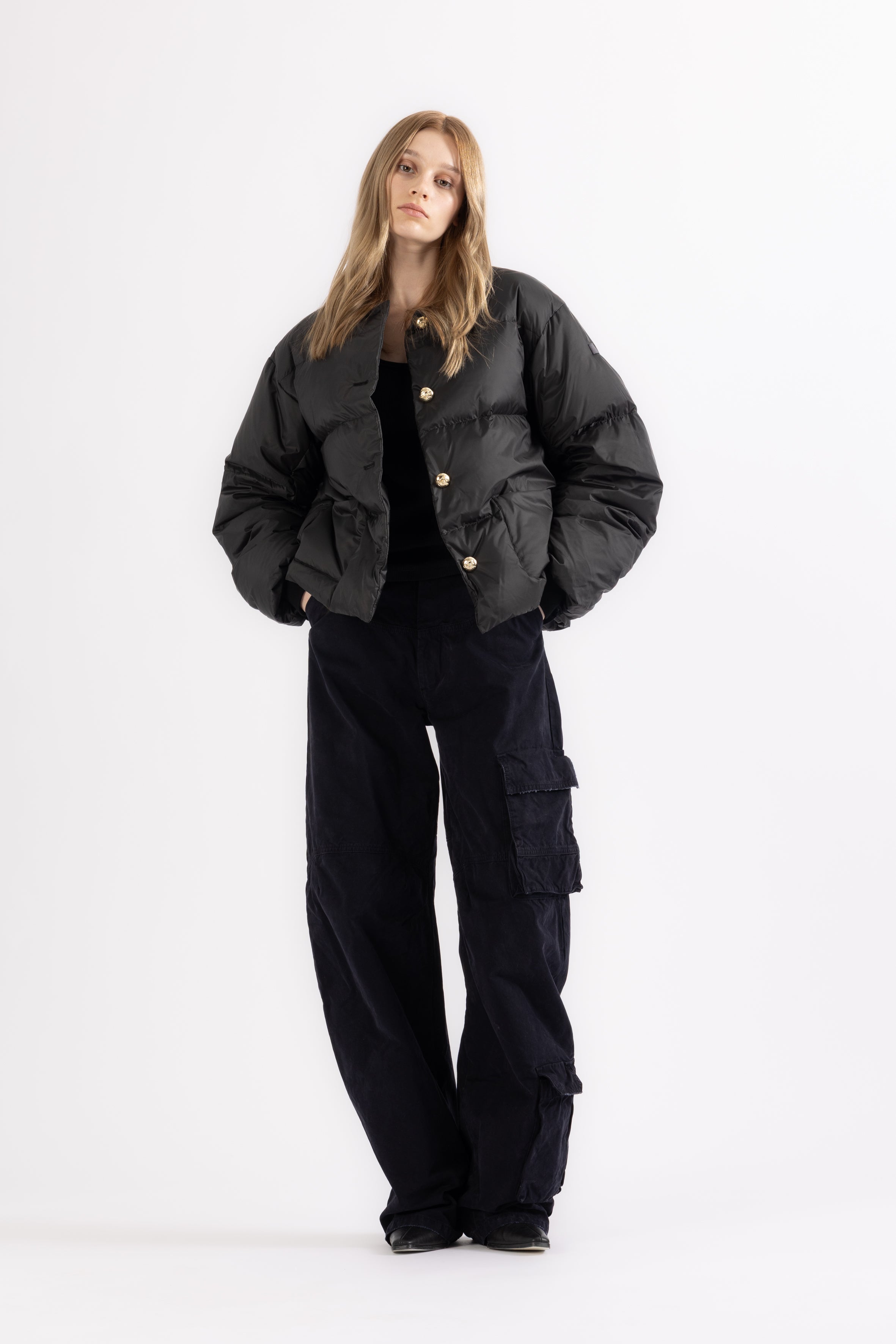 Person wearing a black LEMPELIUS down jacket 278GO with gold buttons and cargo pants on a white background