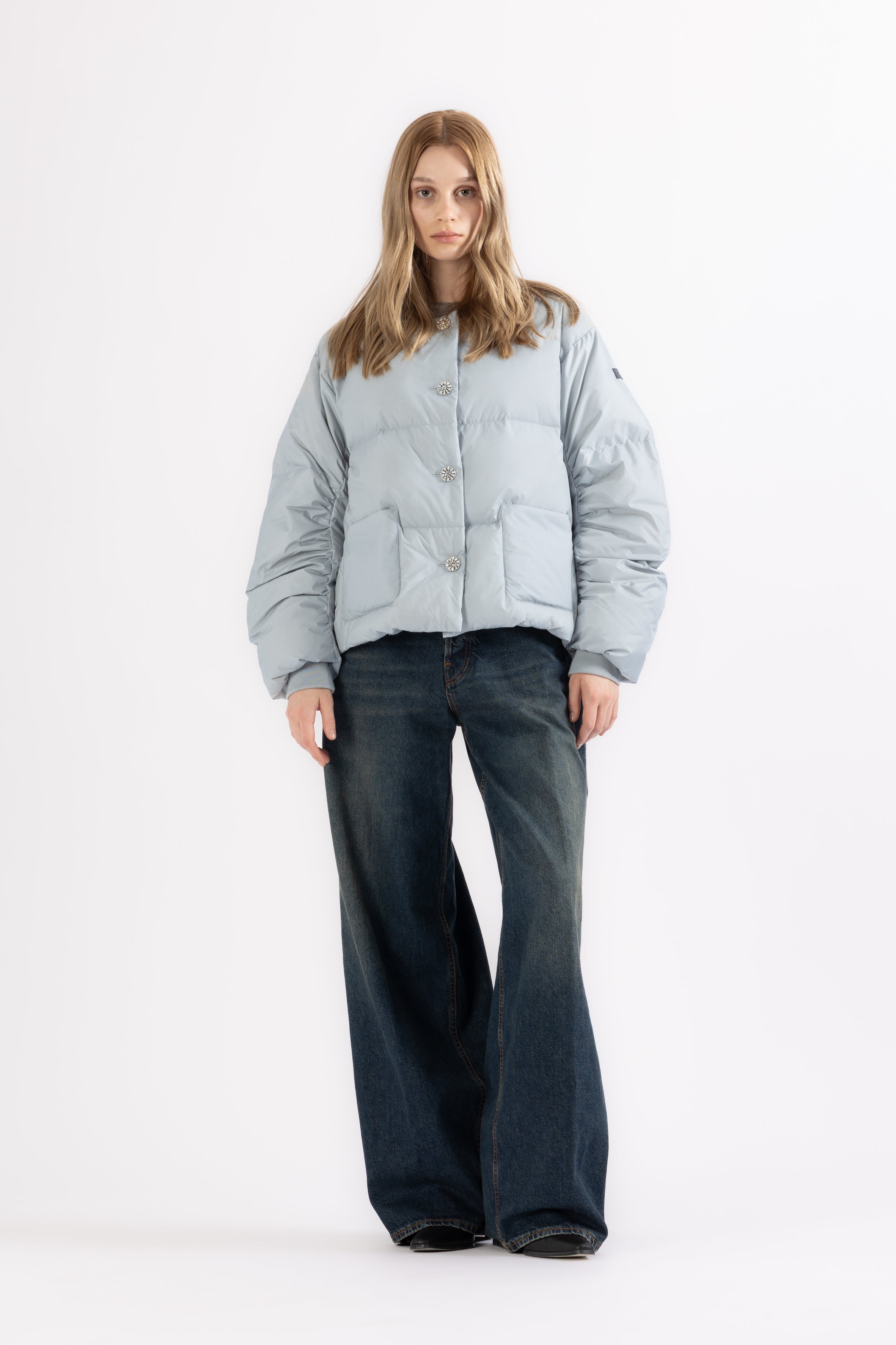 Person wearing a light gray blueish LEMPELIUS puffer jacket with rhinestone buttons and dark jeans on a white background